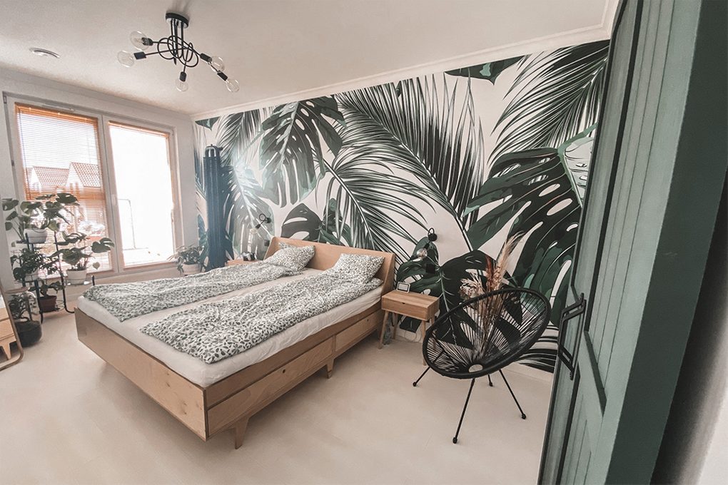 wnętrze sypialni z jasnym drewnianym łóżkiem, dekoracyjną tapetą z motywem roślinnym i czarnym krzesłem ażurowym