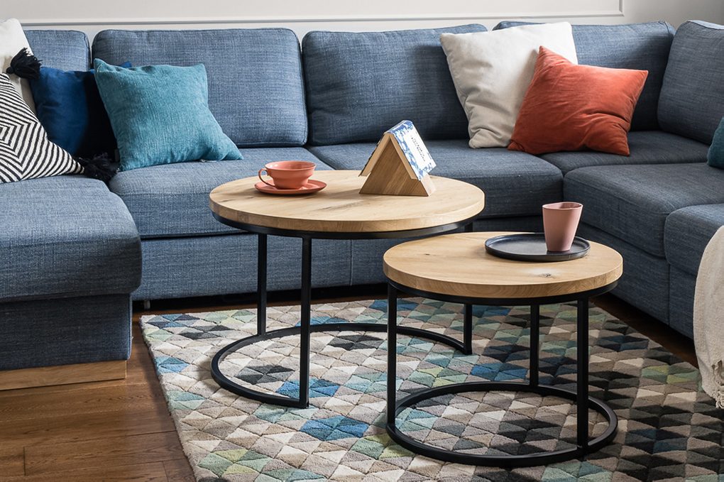 Zestaw dwóch okrągłych stolików kawowych z jasnego drewna i metalu na tle dużej niebieskiej sofy