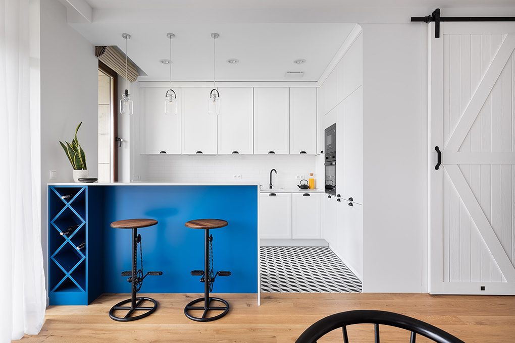 Otwarta kuchnia urządzona w bieli z czarnymi, metalowymi dodatkami oddzielona od salonu barkiem śniadaniowym wykończonym na niebiesko