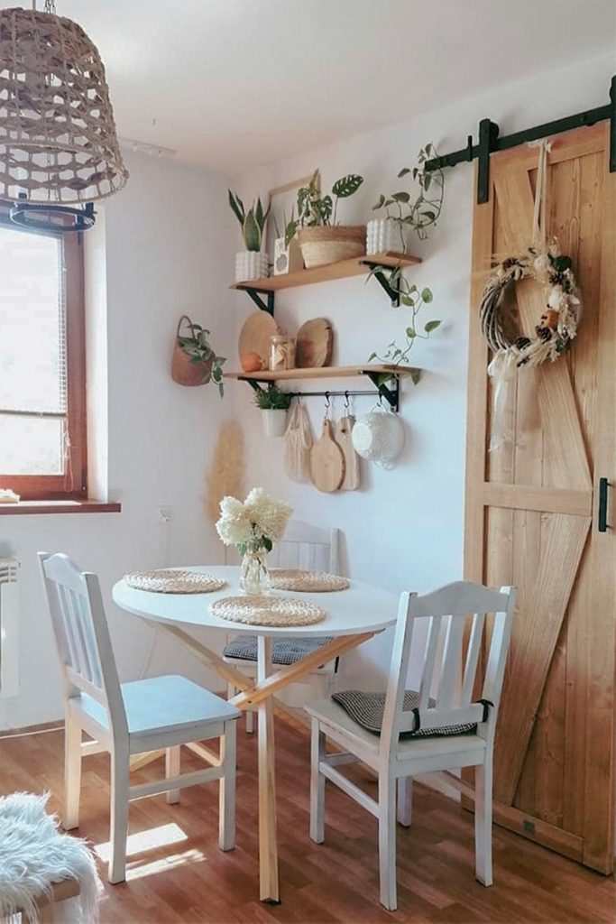 biały okrągły stół z białymi krzesłami stojący przy ścianie, na której wiszą półki z roślinami