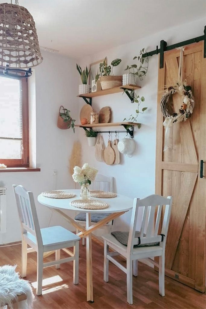 biały okrągły stół z białymi krzesłami stojący przy ścianie, na której wiszą półki z roślinami