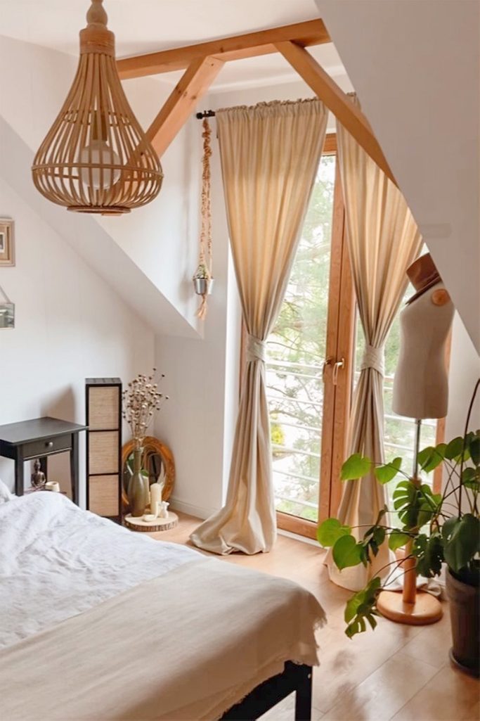 beżowe zasłony, bambusowy żyrandol, drewniane belki przy suficie w naturalnej sypialni