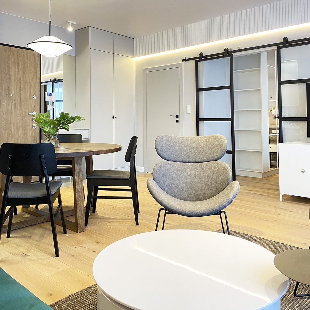 biały okrągły stolik, szare fotele na metalowym stelażu, białe ściany i jasna podłoga w nowoczesnym mieszkaniu