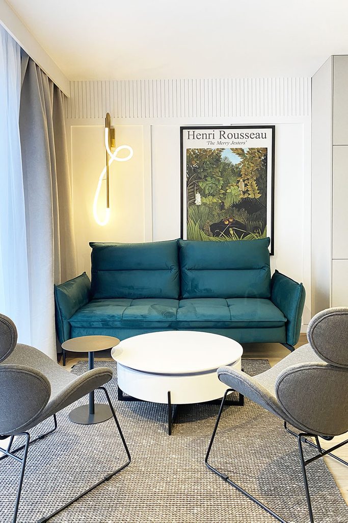 szmaragdowa sofa, biały okrągły stolik kawowy i szare fotele w kąciku wypoczynkowym nowoczesnego mieszkania