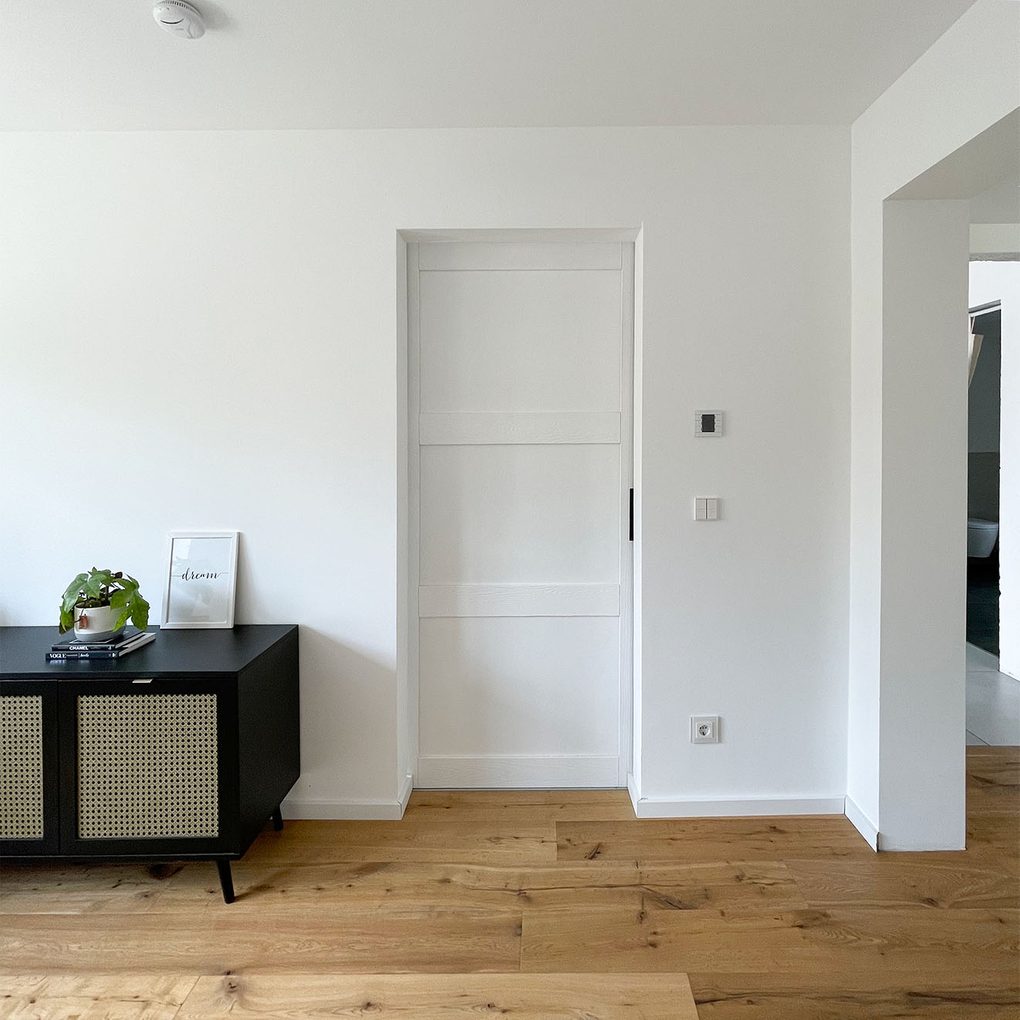 Czarna, niska komoda z ratanem obok białych drzwi w sypialni, na tle białej ściany bez dekoracji