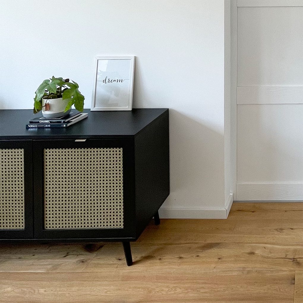 Czarna, niska komoda z ratanem obok białych drzwi w sypialni