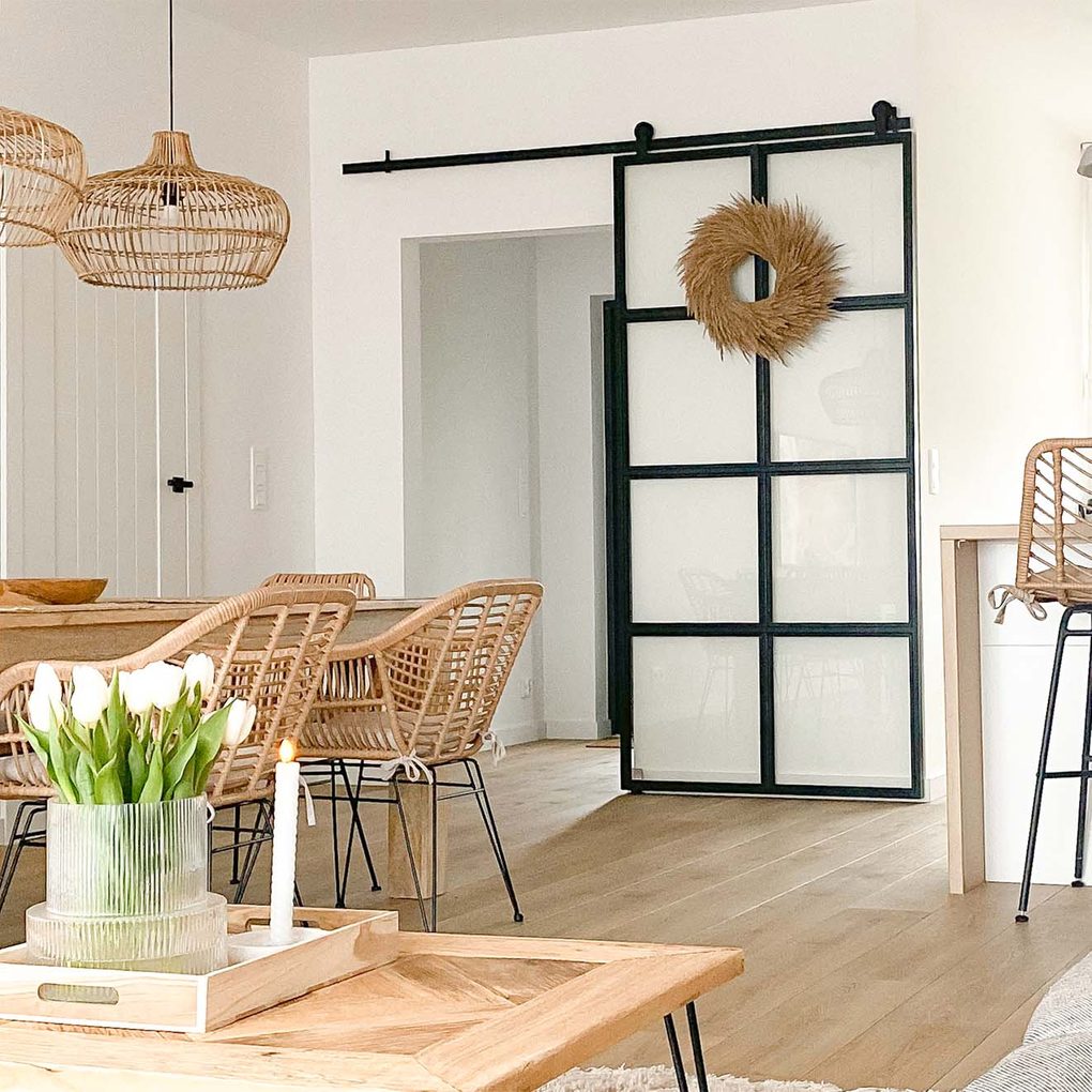 rattanowe krzesła na metalowych nogach, duży drewniany stół, lampy wiszące z wiklinowym abażurem oraz szklane drzwi przesuwne z metalową ramą