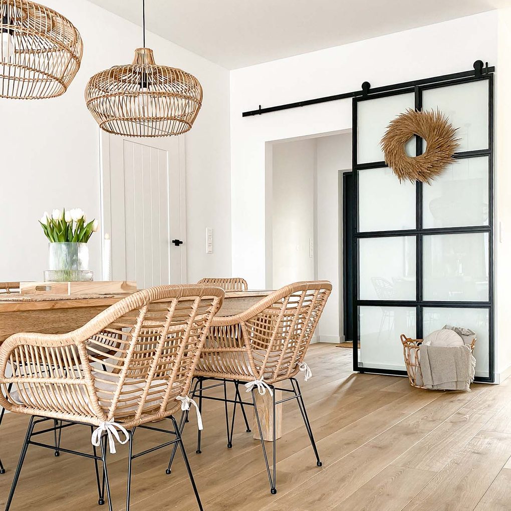 rattanowe krzesła na metalowych nogach, duży drewniany stół, lampy wiszące z wiklinowym abażurem oraz szklane drzwi przesuwne z metalową ramą