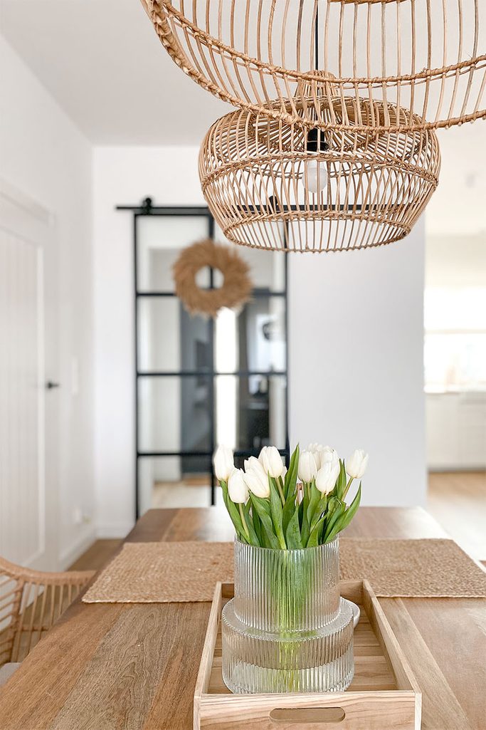 szklany wazon na drewnianej tacy oraz wiklinowe lampy dekorujące salon z białymi ścianami